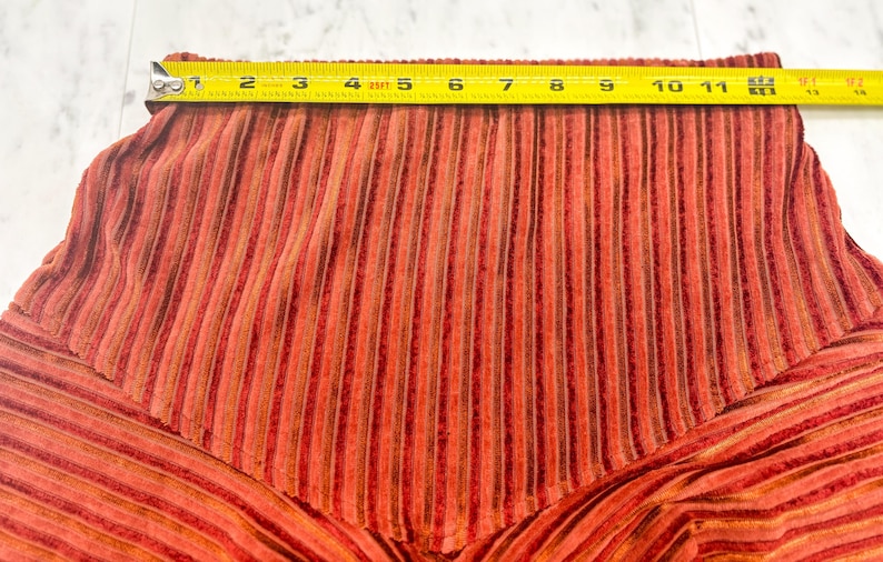 May include: Close-up of a section of orange and brown striped fabric. A yellow tape measure shows the width of the fabric is approximately 11 inches. The fabric has a textured, corduroy-like appearance.
