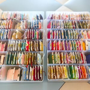 May include: Two clear plastic storage containers filled with numerous embroidery floss skeins wrapped around individual cardboard bobbins. Each bobbin features a small yellow label with a printed number, organized in rows within the compartments.