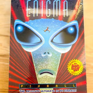May include: A PC game box for "Enigma Pinball" featuring an alien face with glowing blue eyes and the text "The Absolute Ultimate in PC Pinball!" on a red and black background. The box is sealed in plastic.