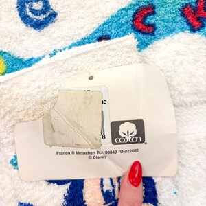 May include: White cotton towel with a colorful cartoon character design. The towel features blue, red, and yellow accents. A tag with the cotton logo and the Disney brand is attached to the towel.