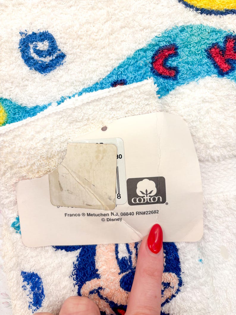 May include: White cotton towel with a colorful cartoon character design. The towel features blue, red, and yellow accents. A tag with the cotton logo and the Disney brand is attached to the towel.