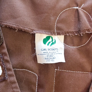May include: Close-up of a brown Girl Scouts uniform with patches and a tag. The tag features the Girl Scouts logo and the words "Girl Scouts Made in U.S.A." The size is Medium (10-12).