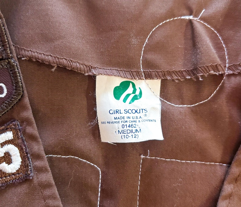 May include: Close-up of a brown Girl Scouts uniform with patches and a tag. The tag features the Girl Scouts logo and the words "Girl Scouts Made in U.S.A." The size is Medium (10-12).