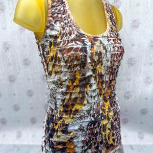 Puede incluir: Una camiseta sin mangas con un diseño texturizado en capas. La camiseta presenta un patrón de blanco, marrón y amarillo, creando una textura visual única. El escote y las correas son de color rosa claro. La parte inferior de la camiseta se combina con un patrón de cuadros marrones y blancos.