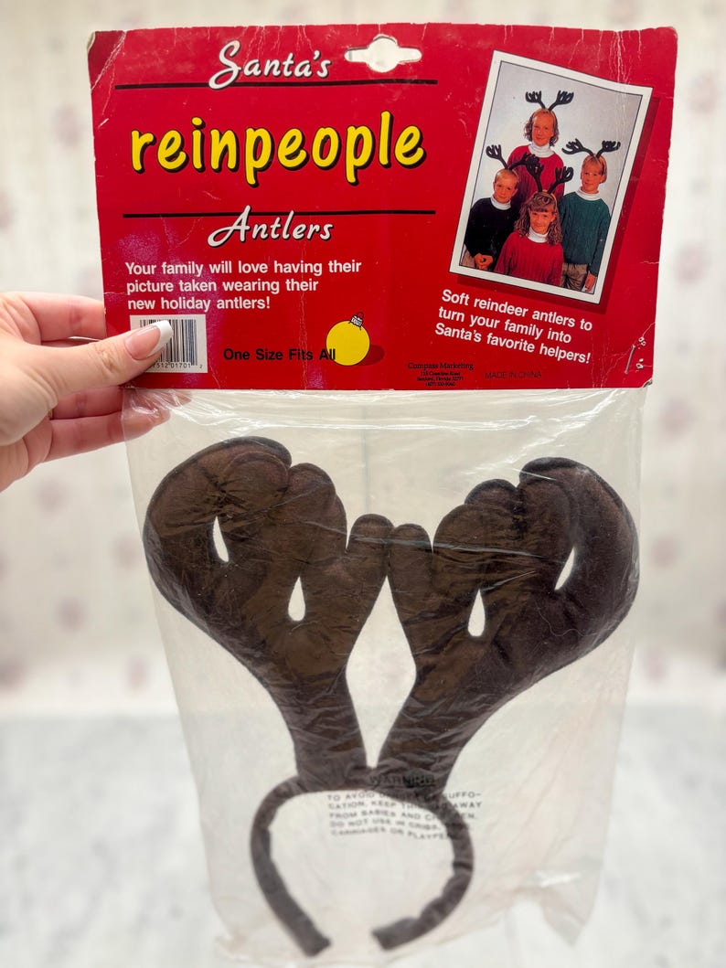 May include: A package of brown reindeer antlers on a headband. The packaging is red with the text "Santa's reinpeople Antlers". The package also includes a photo of children wearing the antlers. One size fits all.