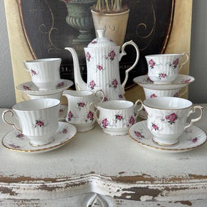 May include: A white tea set with pink floral accents. The set includes a teapot, creamer, sugar bowl, and four teacups and saucers.