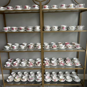 May include: A gold metal shelf unit with five shelves displaying a collection of teacups and saucers with floral patterns. The teacups and saucers are arranged in rows on the shelves.