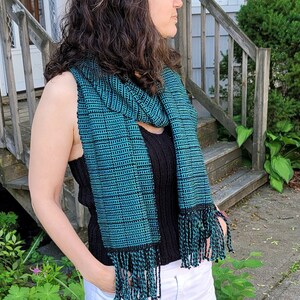 May include: A woman wearing a black top with a teal and black woven scarf, white cropped pants, and white sandals.