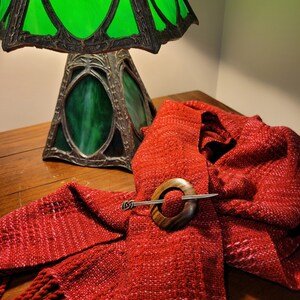 May include: A red woven scarf with a wooden ring and silver pin. The scarf is draped over a wooden table with a green stained glass lamp in the background.