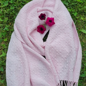 Può includere: Una sciarpa rosa tessuta con una texture delicata e frange. Tre fiori rosa sono posizionati sopra la sciarpa.