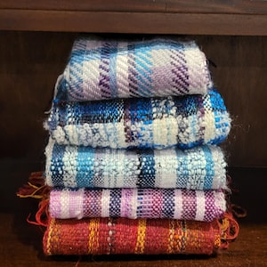Puede incluir: Una pila de cinco cuadrados textiles tejidos a mano en varios colores. El cuadrado superior es azul y morado, seguido de azul, azul claro, rosa y morado, y finalmente, rojo y naranja. Cada cuadrado tiene un patrón tejido único.