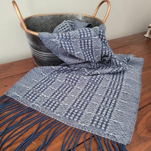 May include: A woven scarf with a blue and white geometric pattern and fringe. The scarf is draped over a wooden surface and partially inside a galvanized metal bucket with gold-colored handles.