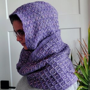 May include: A hooded scarf in shades of purple, with a textured, woven appearance. The scarf is wrapped around the neck and head, with a grid-like pattern created by white threads. The fabric appears soft and warm, suitable for cold weather.