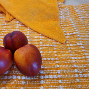 May include: Three ripe nectarines with red and yellow skin rest on a yellow and white patterned tablecloth. A folded yellow cloth is in the background. The image is a close-up shot of the fruit and fabric.