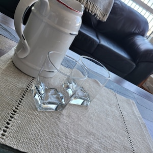 May include: Two clear glass tumblers on a beige placemat with a white pitcher in the background.