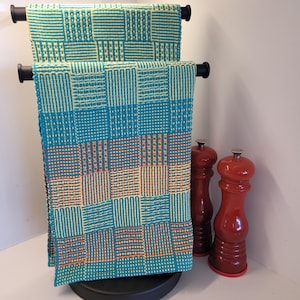 May include: Two teal and yellow woven kitchen towels with a geometric pattern are displayed on a black stand. Two red pepper grinders stand next to the towels. The towels are made of a woven fabric with a grid-like design.