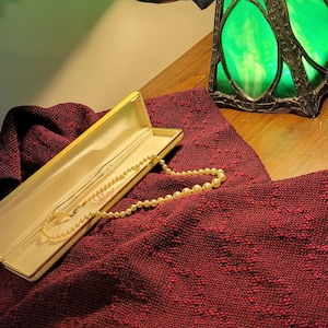 May include: A pearl necklace in a yellow box on a red patterned fabric with a green stained glass lamp in the background.