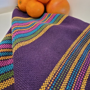Puede incluir: Un textil tejido morado doblado con acentos a rayas coloridas en rosa, amarillo, azul y verde. El textil está drapeado, con una superficie texturizada. Se ven naranjas y pomelos en el fondo.