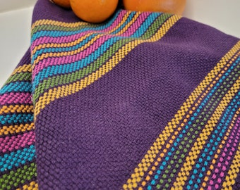 May include: A folded purple woven textile with colorful striped accents in pink, yellow, blue, and green. The textile is draped, with a textured surface. Oranges and grapefruits are visible in the background.