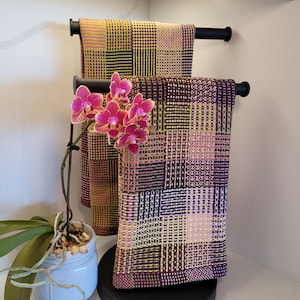 May include: Two black towel racks with a set of two woven towels. The towels are a mix of pink, yellow, and black. A pink orchid plant is in front of the towels.