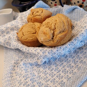 Pode incluir: Três muffins dourados em uma toalha de chá com padrão azul e branco. Os muffins são ligeiramente arredondados e têm uma textura macia e fofa.