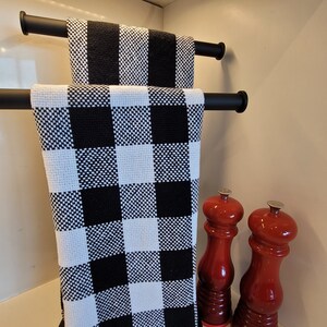 May include: Two black and white checkered kitchen towels hanging on a black towel rack. Two red pepper mills are on the counter below the towels.