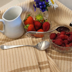May include: A still life arrangement with fresh strawberries and raspberries in glass bowls, a white jug, and a small vase of flowers. A long silver spoon rests on a beige striped cloth.