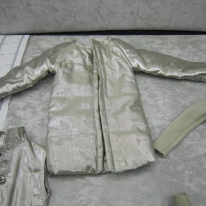 Pu&ograve; includere: Una collezione di abiti per bambole in vari stili e colori. L'immagine presenta una giacca trapuntata argentata, un top senza maniche con un bottone decorativo e una camicia a maniche lunghe. Gli indumenti sono disposti su una superficie grigio chiaro.