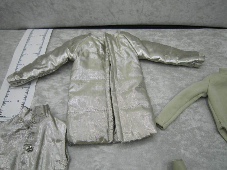Pu&ograve; includere: Una collezione di abiti per bambole in vari stili e colori. L'immagine presenta una giacca trapuntata argentata, un top senza maniche con un bottone decorativo e una camicia a maniche lunghe. Gli indumenti sono disposti su una superficie grigio chiaro.
