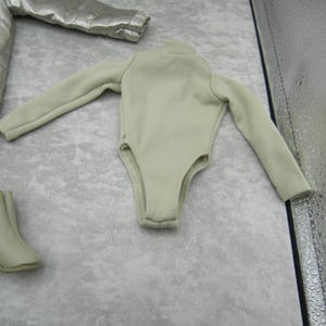 Pu&ograve; includere: Un body beige chiaro con maniche lunghe e collo alto. L'immagine mostra anche una giacca argentata e due paia di stivali beige abbinati. Gli articoli sembrano essere vestiti per bambole.