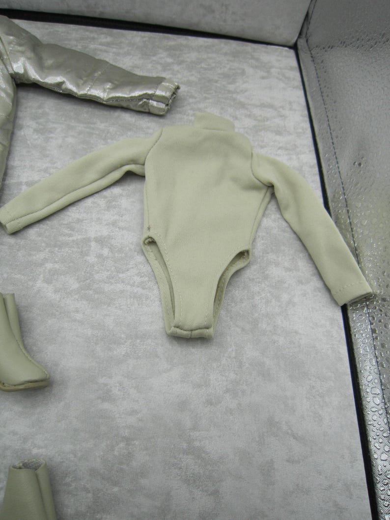 Pu&ograve; includere: Un body beige chiaro con maniche lunghe e collo alto. L'immagine mostra anche una giacca argentata e due paia di stivali beige abbinati. Gli articoli sembrano essere vestiti per bambole.