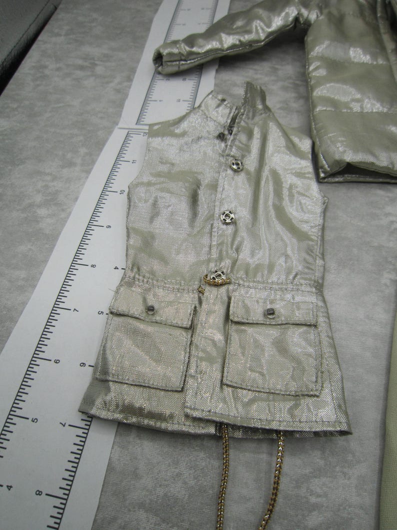 Pu&ograve; includere: Un gilet color argento con due tasche frontali e bottoni decorativi. Il gilet ha un colletto alto e una catena color oro. &Egrave; visibile anche una giacca argentata abbinata.