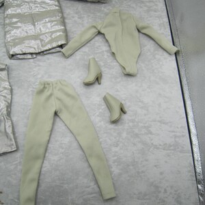 Pu&ograve; includere: Un set di abbigliamento per bambole in tonalit&agrave; neutre. Il set comprende un cappotto argentato trapuntato, un body beige chiaro, pantaloni coordinati e due paia di stivaletti beige con tacco. Gli articoli sono disposti su una superficie grigio chiaro.