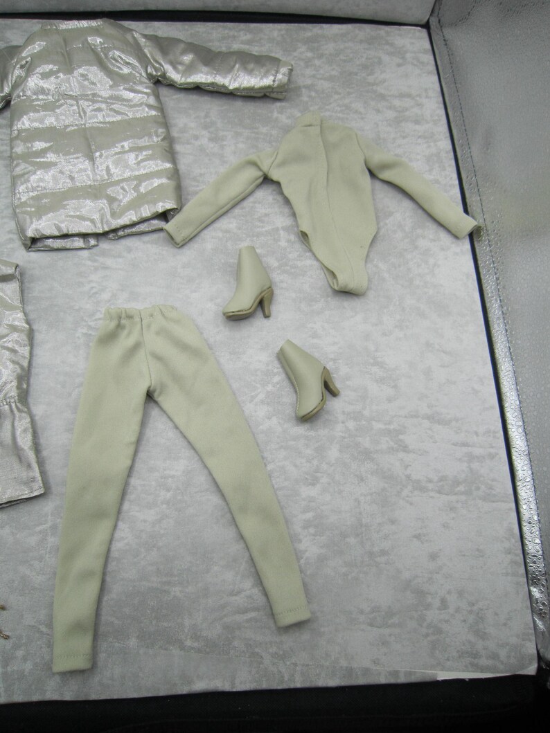 Pu&ograve; includere: Un set di abbigliamento per bambole in tonalit&agrave; neutre. Il set comprende un cappotto argentato trapuntato, un body beige chiaro, pantaloni coordinati e due paia di stivaletti beige con tacco. Gli articoli sono disposti su una superficie grigio chiaro.