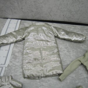Pu&ograve; includere: Una giacca trapuntata color argento con maniche lunghe. La giacca &egrave; realizzata in un tessuto lucido e metallico e ha un taglio lungo e dritto. L'immagine mostra anche una camicia a maniche lunghe verde chiaro e un piccolo pezzo di giacca argentata.