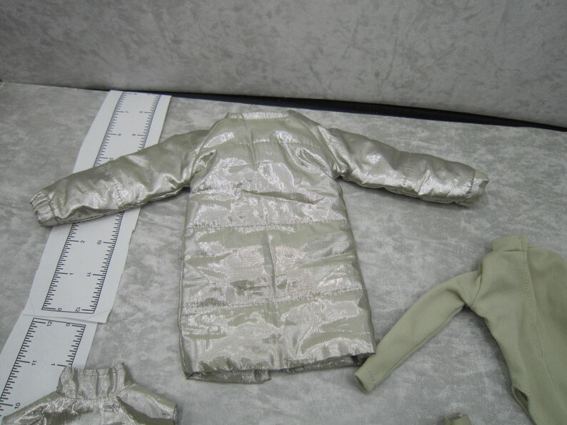 Pu&ograve; includere: Una giacca trapuntata color argento con maniche lunghe. La giacca &egrave; realizzata in un tessuto lucido e metallico e ha un taglio lungo e dritto. L'immagine mostra anche una camicia a maniche lunghe verde chiaro e un piccolo pezzo di giacca argentata.