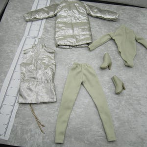 Pu&ograve; includere: Un set di moda per bambole con giacca puffer metallizzata argentata, top senza maniche, leggings, body e due paia di stivaletti. L'abbigliamento &egrave; di colore beige chiaro. Il set &egrave; disposto su una superficie grigio chiaro.