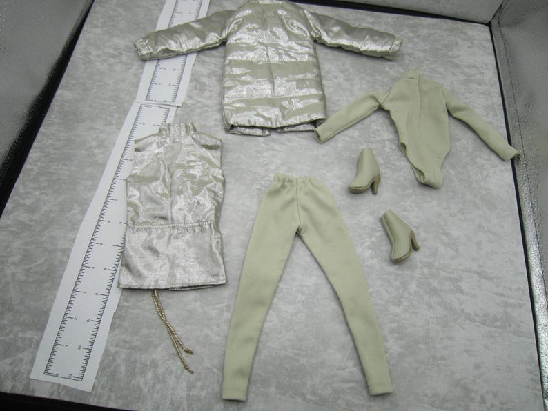 Pu&ograve; includere: Un set di moda per bambole con giacca puffer metallizzata argentata, top senza maniche, leggings, body e due paia di stivaletti. L'abbigliamento &egrave; di colore beige chiaro. Il set &egrave; disposto su una superficie grigio chiaro.