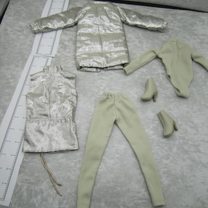 Pu&ograve; includere: Un set di moda per bambole con giacca trapuntata argentata, top all'americana abbinato, pantaloni, body e due paia di stivaletti con tacco. L'abbigliamento &egrave; di colore chiaro e neutro, disposto su una superficie chiara.
