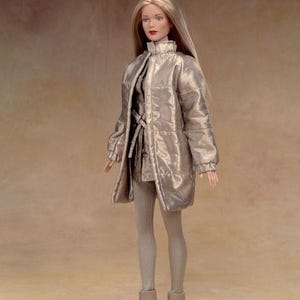 Pu&ograve; includere: Bambola alla moda con lunghi capelli biondi, indossa un cappotto metallizzato argentato, un vestito abbinato, leggings grigi e stivaletti grigi. La bambola ha il rossetto rosso e posa su uno sfondo neutro.