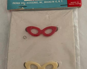 Può includere: Un pacchetto di due paia di occhiali di plastica per bambole. Un paio è rosso e l'altro è color crema. La confezione è etichettata "Premier Product, Toyland's Glamour Stylists for Dolls, No. 1028, Size Bobby, Premier Doll Accessories, Inc., Brooklyn 15, N.Y., Made in Hong Kong".