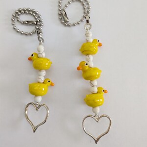 May include: Two beaded charm necklaces. Each necklace features three yellow duck charms with orange beaks, white beads, and a silver heart pendant. The necklaces hang from a silver ball chain.