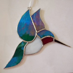 Op de afbeelding: Een glazen kolibrie-ornament met blauwe, groene, witte en rode glasstukken. De kolibrie is in vlucht met zijn vleugels uitgespreid.