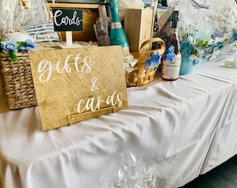以下が含まれることがあります： 「gifts & cards」と白文字で書かれた木製の看板が、籐のバスケット、ワインボトル、その他のギフトが置かれたテーブルに置かれています。