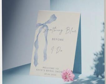 以下が含まれることがあります： 「Something Blue Before I Do」のテキストと水色のリボンデザインが施されたクリーム色のサイン。ピンクの花がサインの基部にあります。サインはKatie's Bridal Show用で、日付は09.02.2023です。