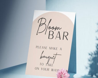 以下が含まれることがあります： 桃色の背景に円形で「Bloom Bar」と書かれた看板。下のテキストには「お持ち帰り用のブーケを作ってください！」と書いてあります。