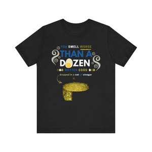 May include: Black t-shirt with a humorous graphic. The text reads "YOU SMELL WORSE THAN A DOZEN ROTTEN EGGS dropped in a vat of vinegar." The design includes a cartoon egg and stylized swirls.
