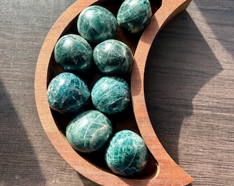 May include: A crescent moon-shaped wooden bowl containing eight polished, rounded green apatite stones. The stones exhibit deep teal and forest green hues with natural white veining patterns throughout their smooth, glossy surfaces.