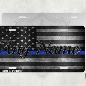 May include: A black and white American flag with a blue stripe across the middle. The text "Any Name" is written in black across the blue stripe. The flag is on a metal license plate.