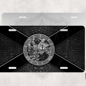 May include: Black and white license plate featuring the Great Seal of the State of Florida with a crocodile skin background. The seal depicts a woman holding a palm tree branch, a steamboat, and the words "In God We Trust".